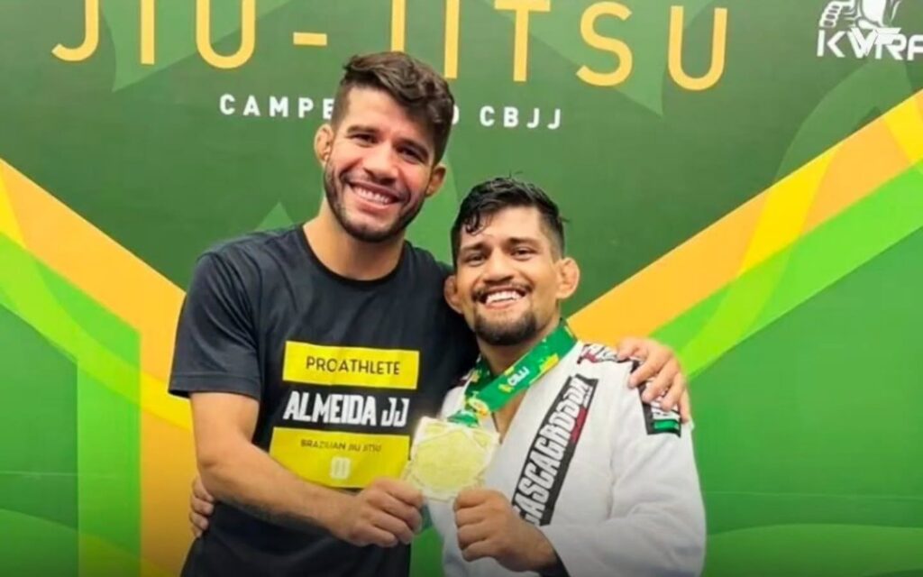 Caio Almeida, in an Almeida JJ shirt, hugs Cleber Clandestino, in a white gi with a gold medal, on a CBJJ podium