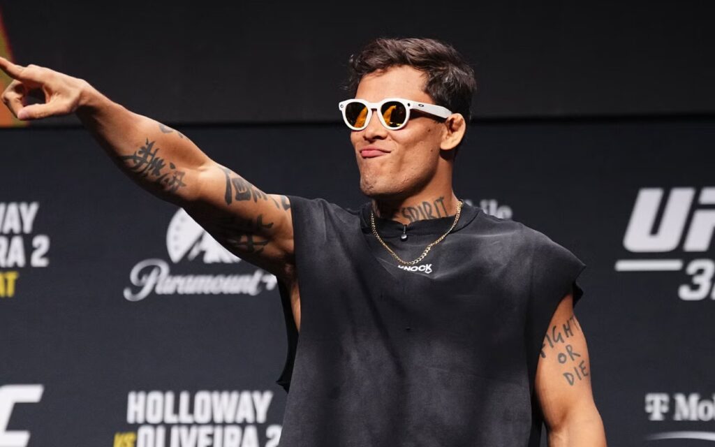 Caio Borralho de óculos escuros brancos e regata preta apontando na coletiva do UFC 326