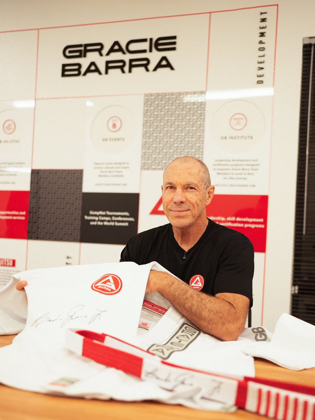 Carlos Gracie Jr. sentado à mesa com kimonos, diante de um painel da Gracie Barra