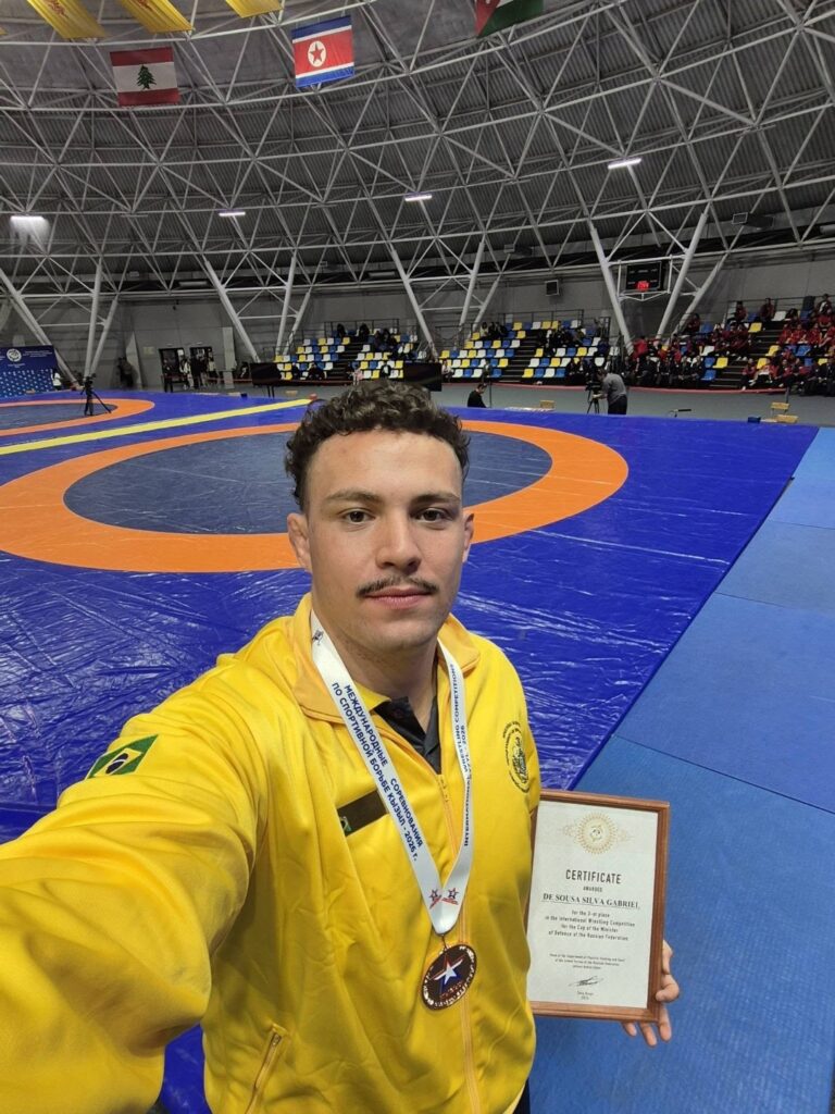 Brasil leva dois bronzes no wrestling na Rússia 1 Gabriel Silva, de agasalho amarelo do Brasil, mostra a medalha de bronze e o diploma no ginásio em Kyzyl
