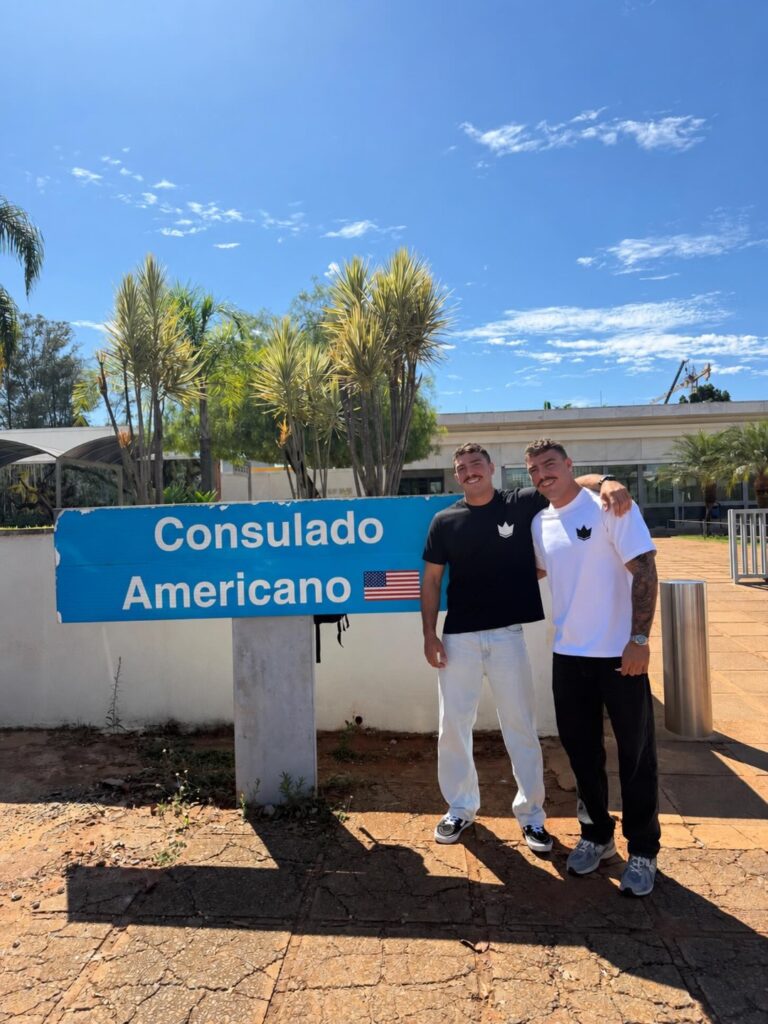 Davi e Matheus Vetoraci posam abraçados ao lado da placa do Consulado Americano