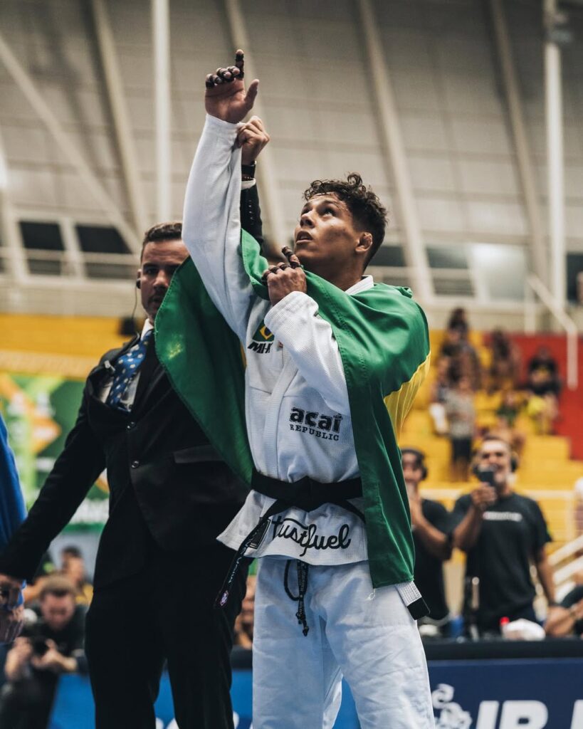 Mica Galvão aponta para o alto, de kimono branco e com uma bandeira nos ombros, ao lado do árbitro